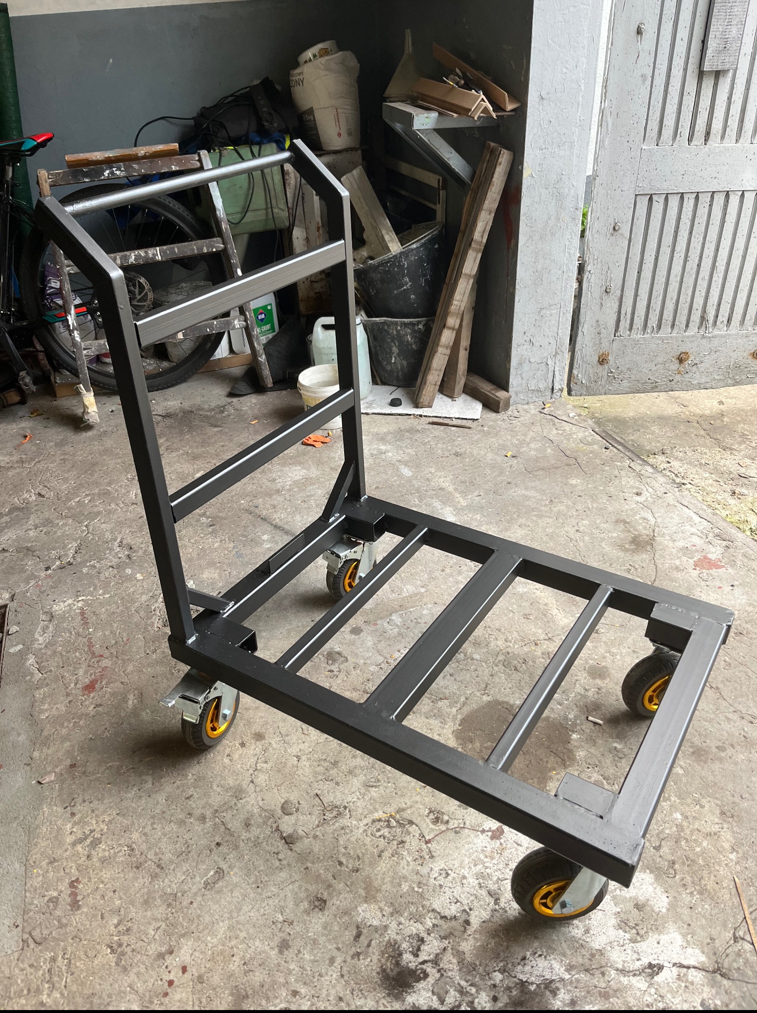 Wózek transportowy ze stali, spawany, malowany proszkowo na czarno, z czterema kołami. Wykonany przez ślusarza z Częstochowy. Widoczne spawy i elementy konstrukcyjne.