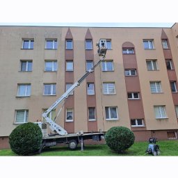 P.u.dziuniek - Praca na wysokościach: podnośnik koszowy przy bloku, pracownik w koszu. Częściowo umyta elewacja w odcieniach beżu i brązu. Widoczna myjka ciśnieniowa na trawniku.