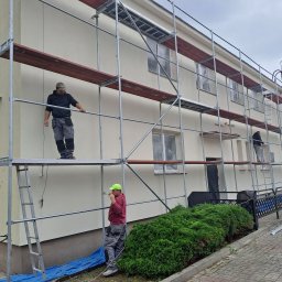 P.u.dziuniek - Dwóch pracowników na rusztowaniu maluje elewację budynku. Okna zabezpieczone folią, widoczny krzew. Jeden z pracowników maluje pędzlem, drugi stoi na rusztowaniu.