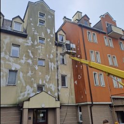 P.u.dziuniek - Mycie elewacji budynku z podnośnika koszowego. Widoczne ślady po czyszczeniu, fragment żółtego ramienia podnośnika i pracownik w dolnym rogu kadru.