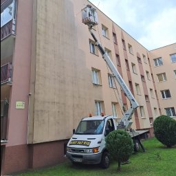 P.u.dziuniek - Pracownik na podnośniku maluje elewację bloku. Częściowo pomalowana ściana, widoczny numer telefonu na samochodzie. Ujęcie z dołu, pochmurne niebo.