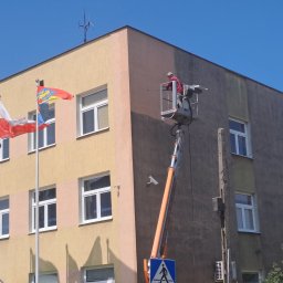 P.u.dziuniek - Pracownik na podnośniku czyści elewację budynku. Widoczne flagi, okna i fragmenty elewacji przed i po czyszczeniu. Ujęcie z dołu, błękitne niebo w tle.