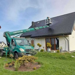 P.u.dziuniek - Pracownik na podnośniku koszowym czyści ciemny dach domu jednorodzinnego. Widoczny zielony trawnik i ozdobne krzewy. Błękitne niebo w tle.