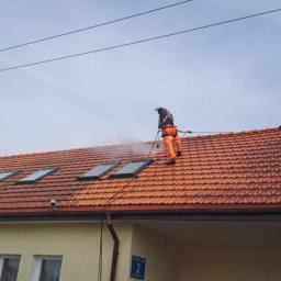 Pracownik w pomarańczowym kombinezonie czyści dach z czerwonej dachówki za pomocą myjki ciśnieniowej. Widoczne okna dachowe i fragment budynku z adresem.