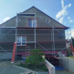 Dom w rusztowaniach podczas prac elewacyjnych. Okna zabezpieczone folią, widoczni pracownicy. Prace w toku, zieleń w tle.