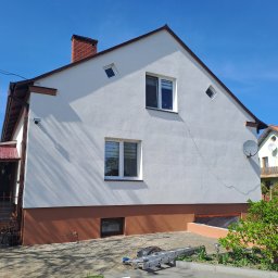 Odnowiona elewacja domu jednorodzinnego w słoneczny dzień. Biała ściana z brązowym cokołem, widoczne okna, dach i drabina oparta o ścianę. Czyste, świeże wykończenie.