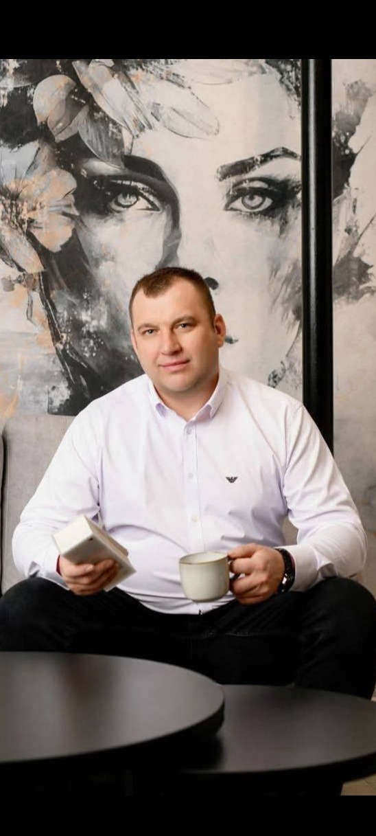 Mężczyzna w białej koszuli siedzi na kanapie z kubkiem i pudełkiem na tle ściany z abstrakcyjnym malowidłem twarzy kobiety. Ujęcie z bliska, naturalne światło.