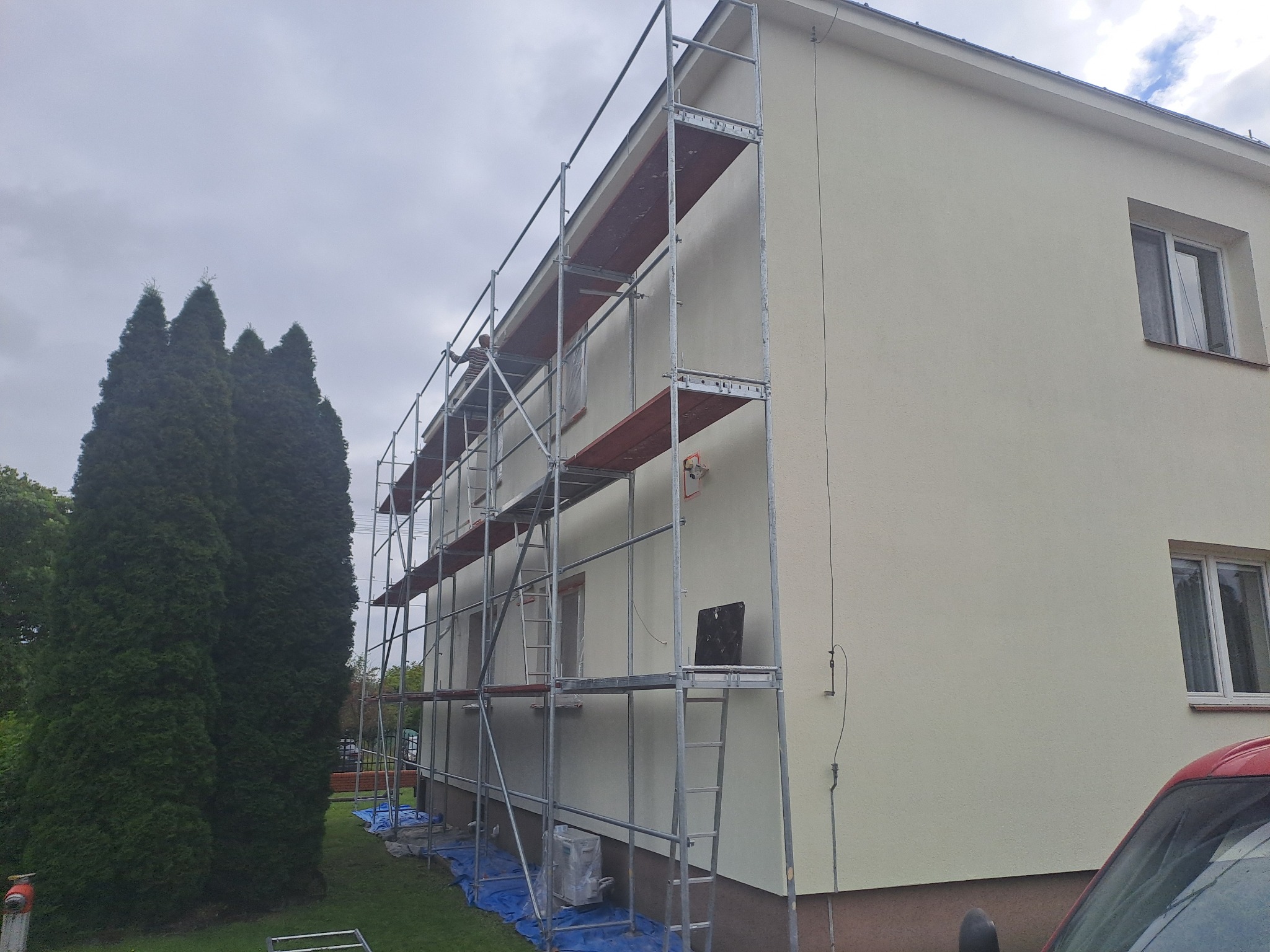 Elewacja domu w trakcie malowania, widoczne rusztowania i zabezpieczenia. Świeżo pomalowana ściana w jasnym odcieniu, zieleń drzew w tle, pochmurne niebo.