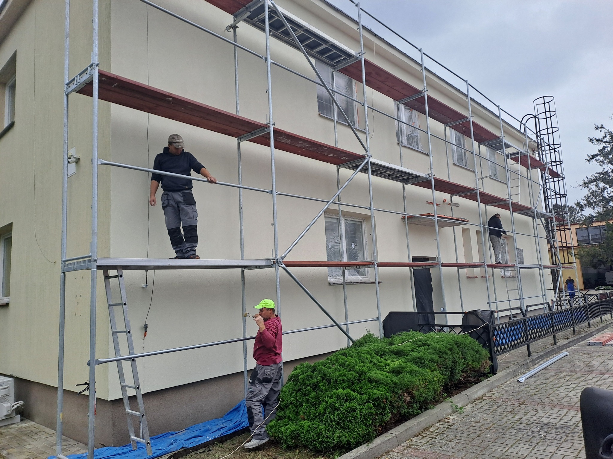 Dwóch pracowników na rusztowaniu maluje elewację budynku. Okna zabezpieczone folią, widoczny krzew. Jeden z pracowników maluje pędzlem, drugi stoi na rusztowaniu.