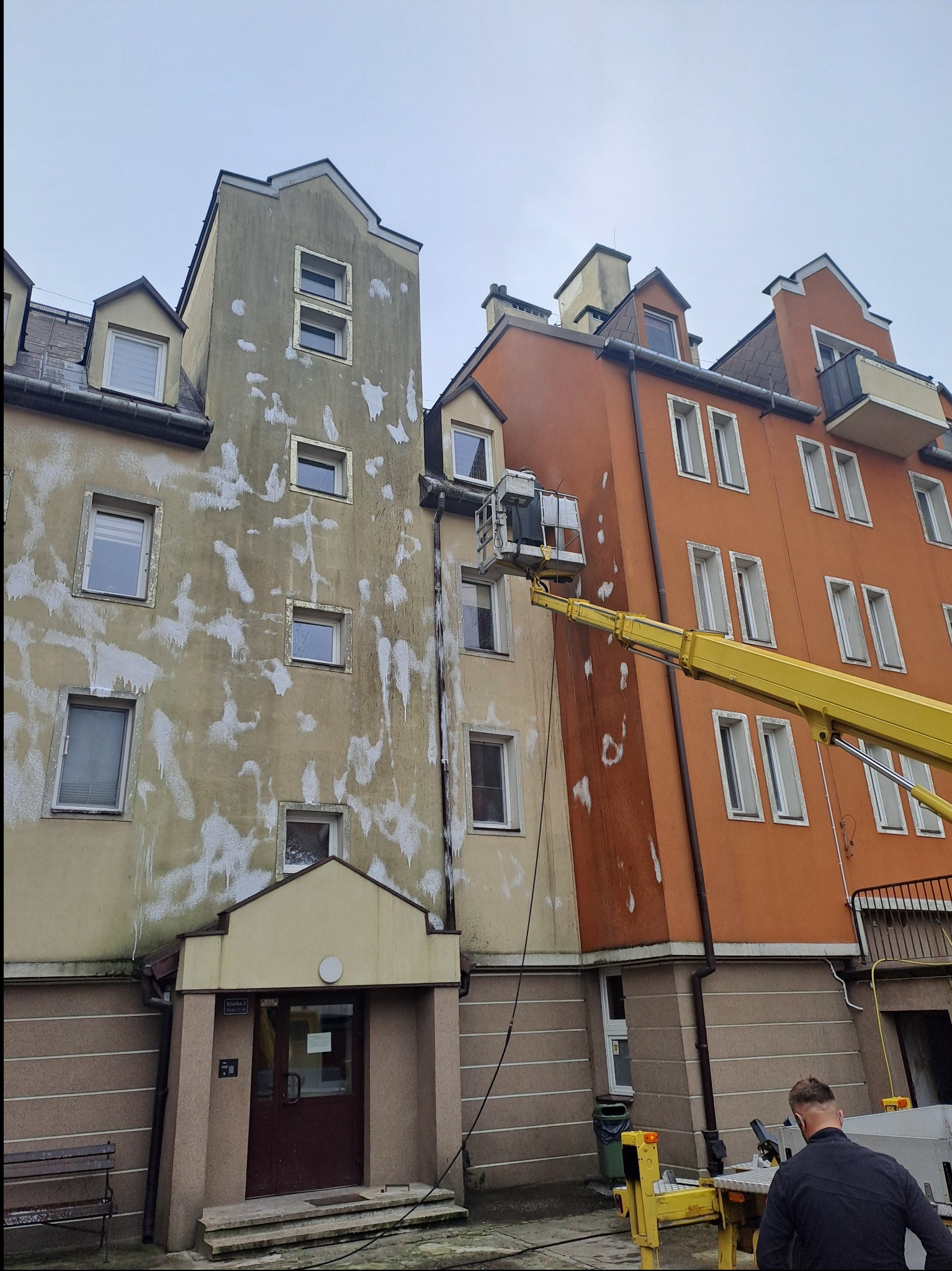 Mycie elewacji budynku z podnośnika koszowego. Widoczne ślady po czyszczeniu, fragment żółtego ramienia podnośnika i pracownik w dolnym rogu kadru.