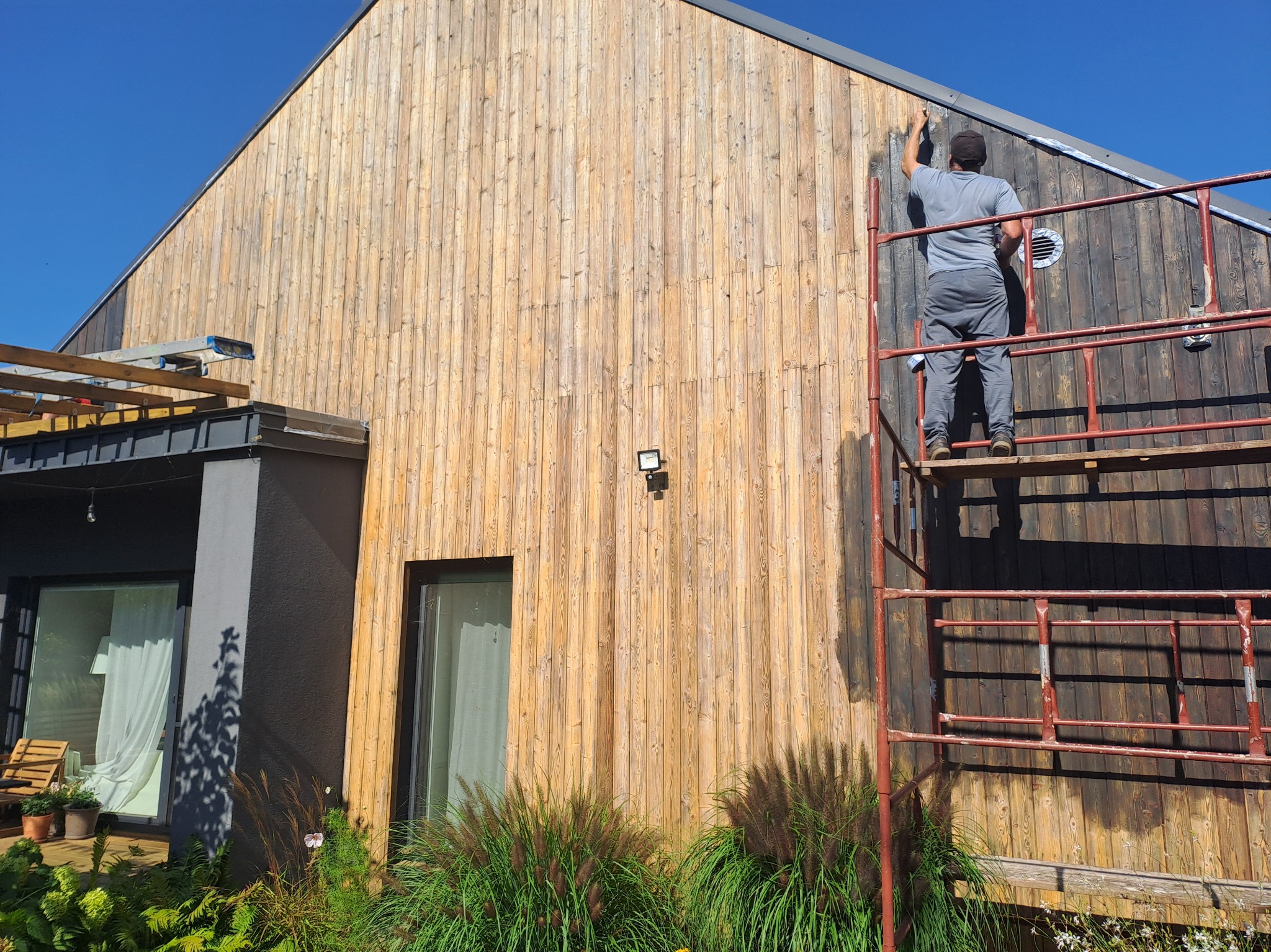 Malowanie drewnianej elewacji domu z rusztowania. Część ściany pomalowana na ciemny kolor, widoczne rośliny i okno. Praca w toku, słoneczny dzień.
