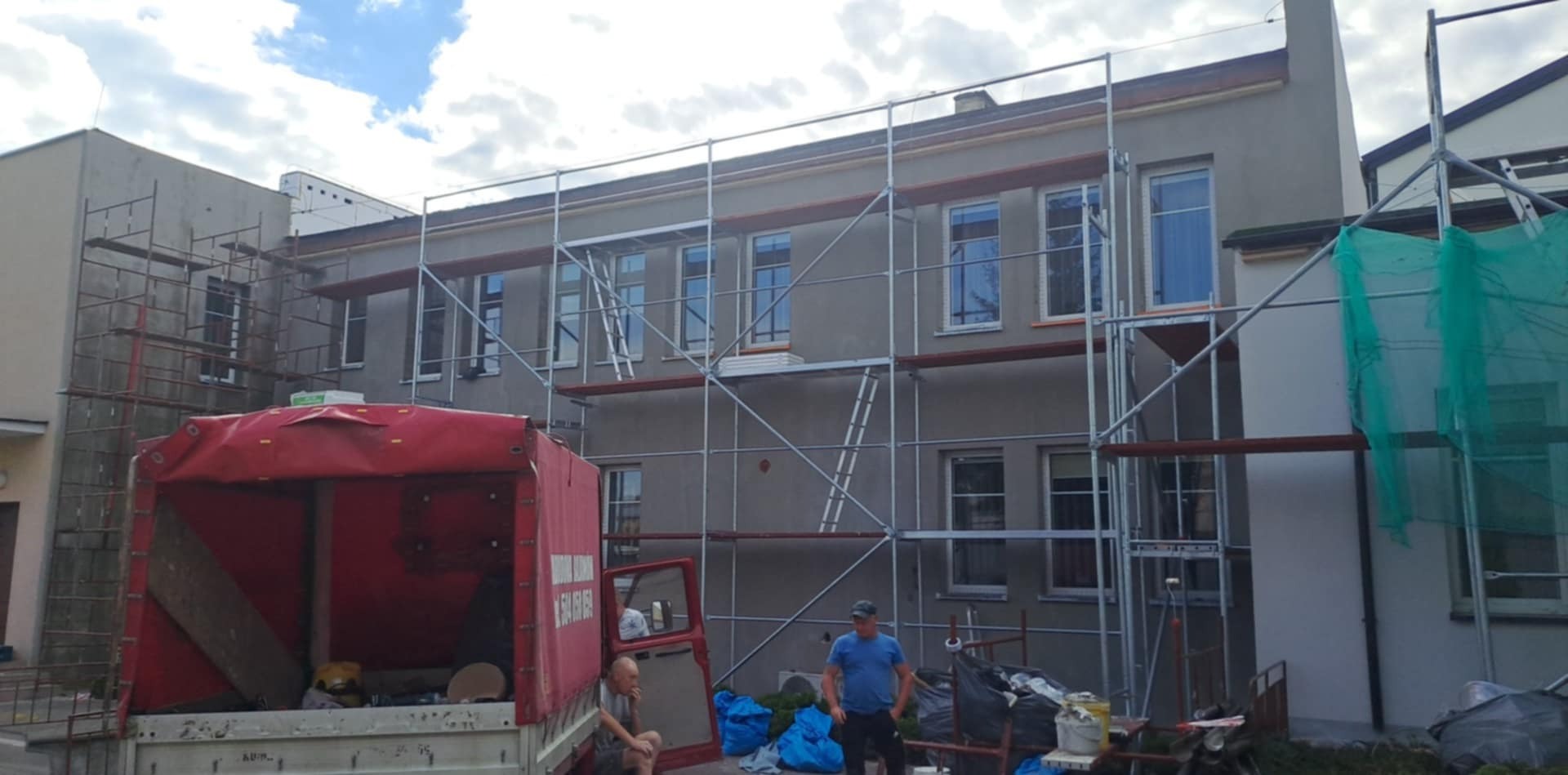 Budynek w trakcie prac elewacyjnych, otoczony rusztowaniami. Widoczny samochód dostawczy i osoby. Świeżo nałożony tynk w szarym odcieniu. Prace w toku.