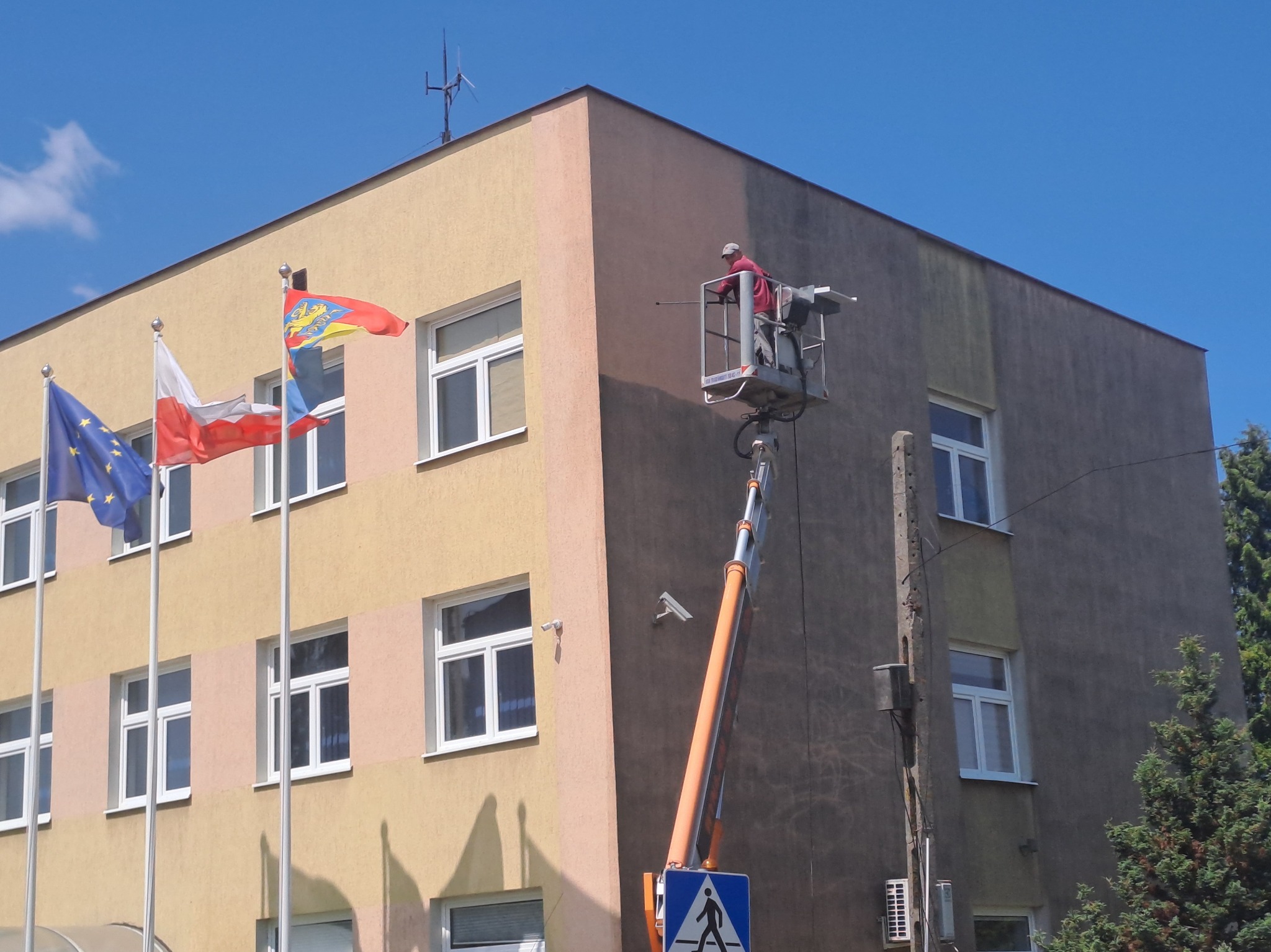 Pracownik na podnośniku czyści elewację budynku. Widoczne flagi, okna i fragmenty elewacji przed i po czyszczeniu. Ujęcie z dołu, błękitne niebo w tle.