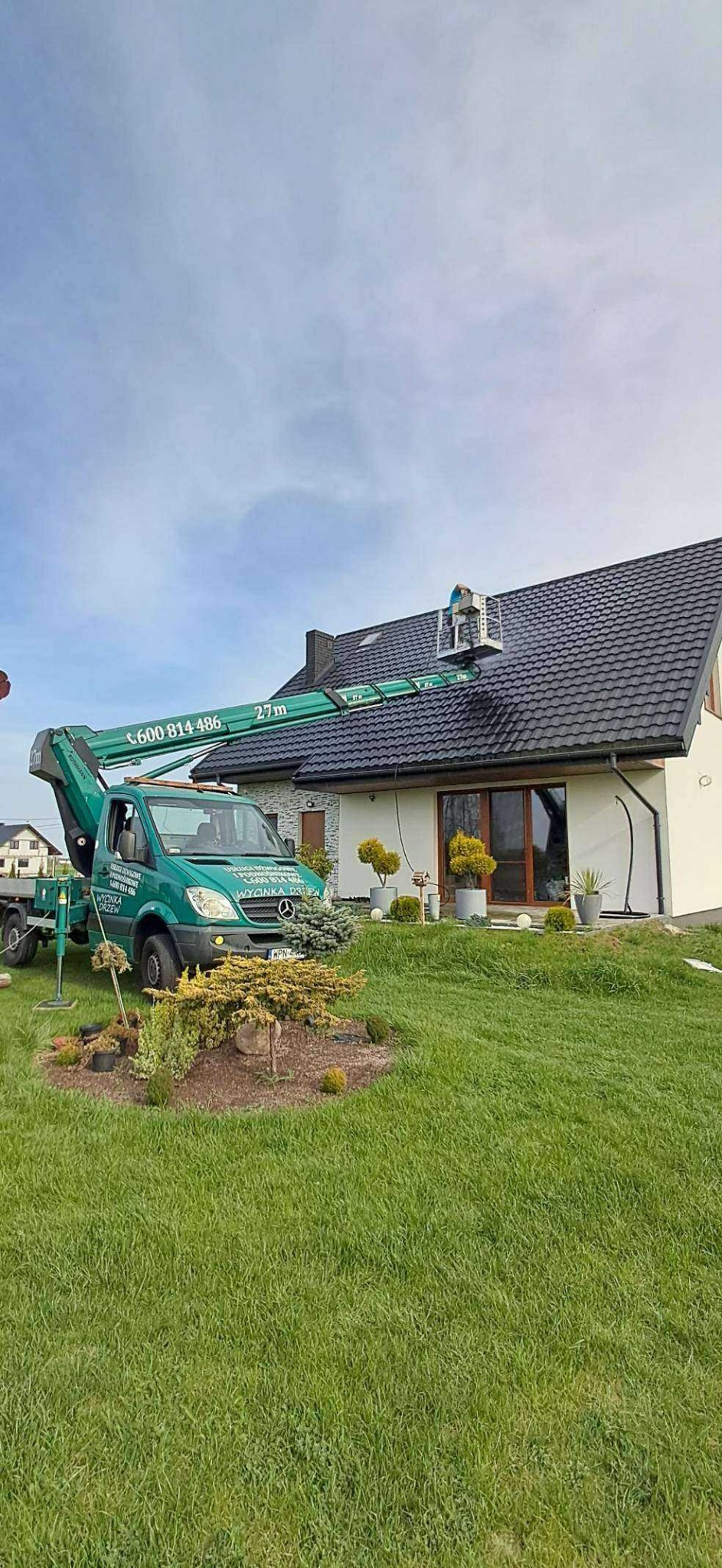 Pracownik na podnośniku koszowym czyści ciemny dach domu jednorodzinnego. Widoczny zielony trawnik i ozdobne krzewy. Błękitne niebo w tle.