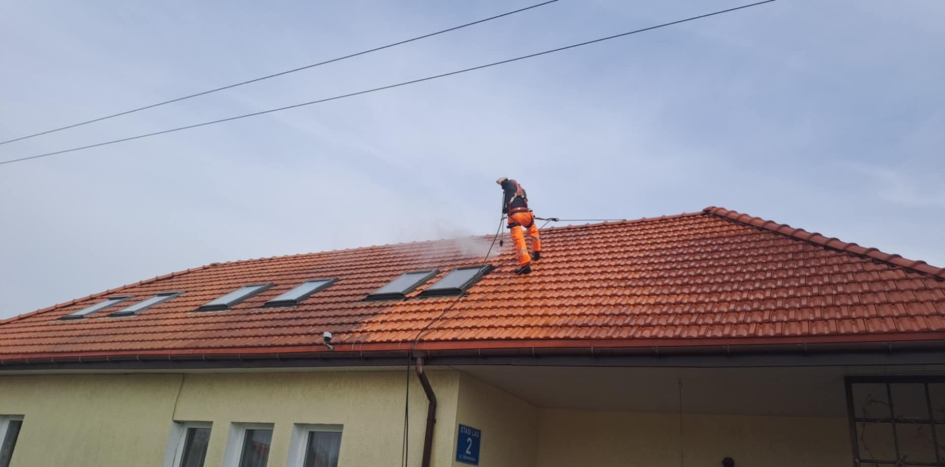 Pracownik w pomarańczowym kombinezonie czyści dach z czerwonej dachówki za pomocą myjki ciśnieniowej. Widoczne okna dachowe i fragment budynku z adresem.