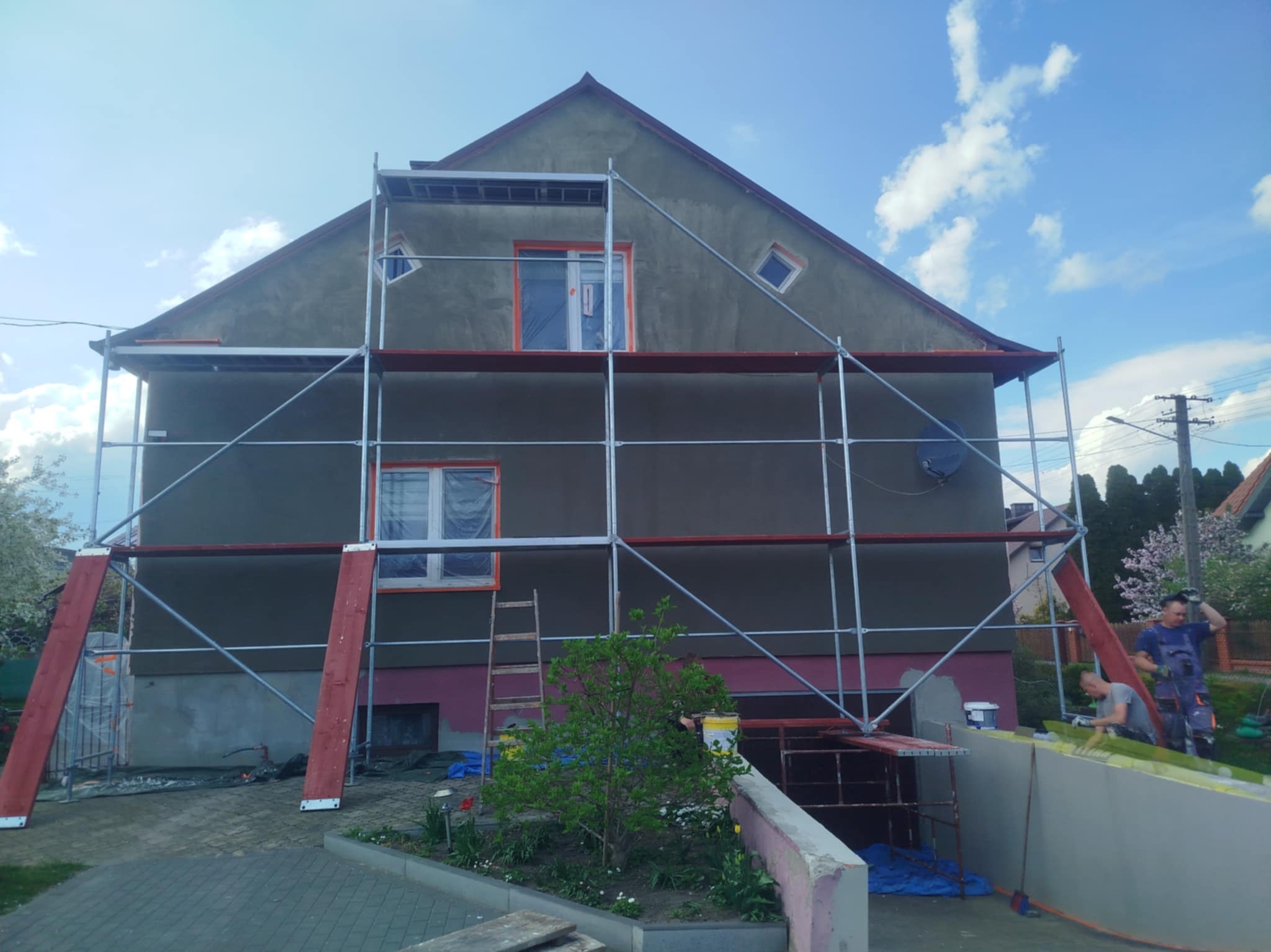 Dom w rusztowaniach podczas prac elewacyjnych. Okna zabezpieczone folią, widoczni pracownicy. Prace w toku, zieleń w tle.