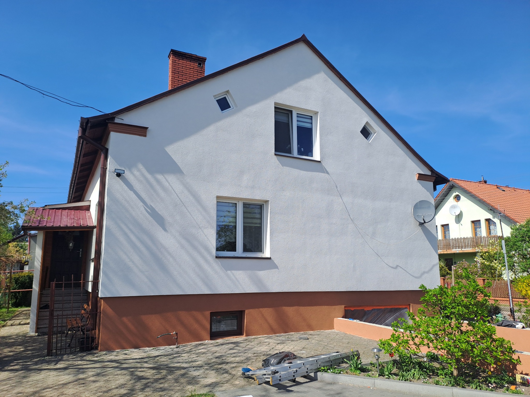 Odnowiona elewacja domu jednorodzinnego w słoneczny dzień. Biała ściana z brązowym cokołem, widoczne okna, dach i drabina oparta o ścianę. Czyste, świeże wykończenie.