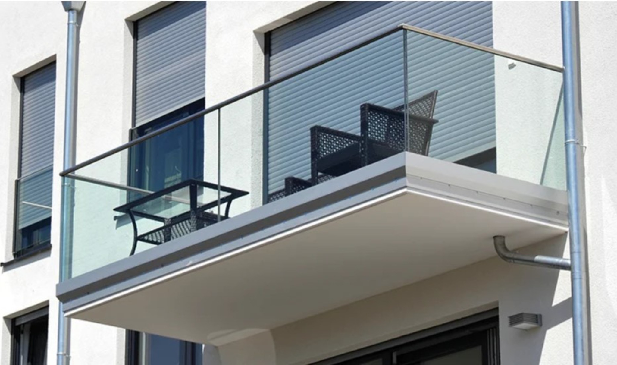 Nowoczesny balkon z szklaną balustradą, wyposażony w czarne meble ogrodowe. Widoczne rolety zewnętrzne na oknach i rynna odprowadzająca wodę. Minimalistyczny design.