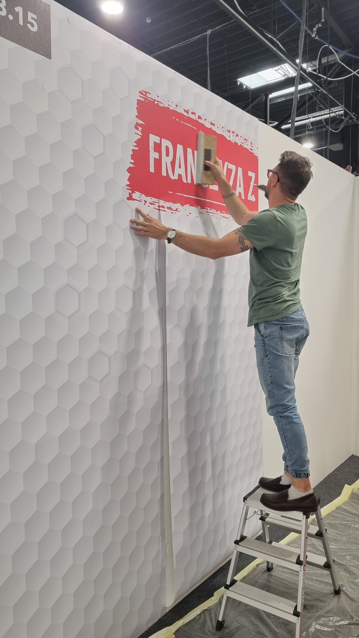 Montaż folii z logo 'FRAN VZA Z' na ścianie z heksagonalną fakturą. Mężczyzna na drabinie dociska folię raklą. Widoczne detale: zegarek, tatuaż na ramieniu.