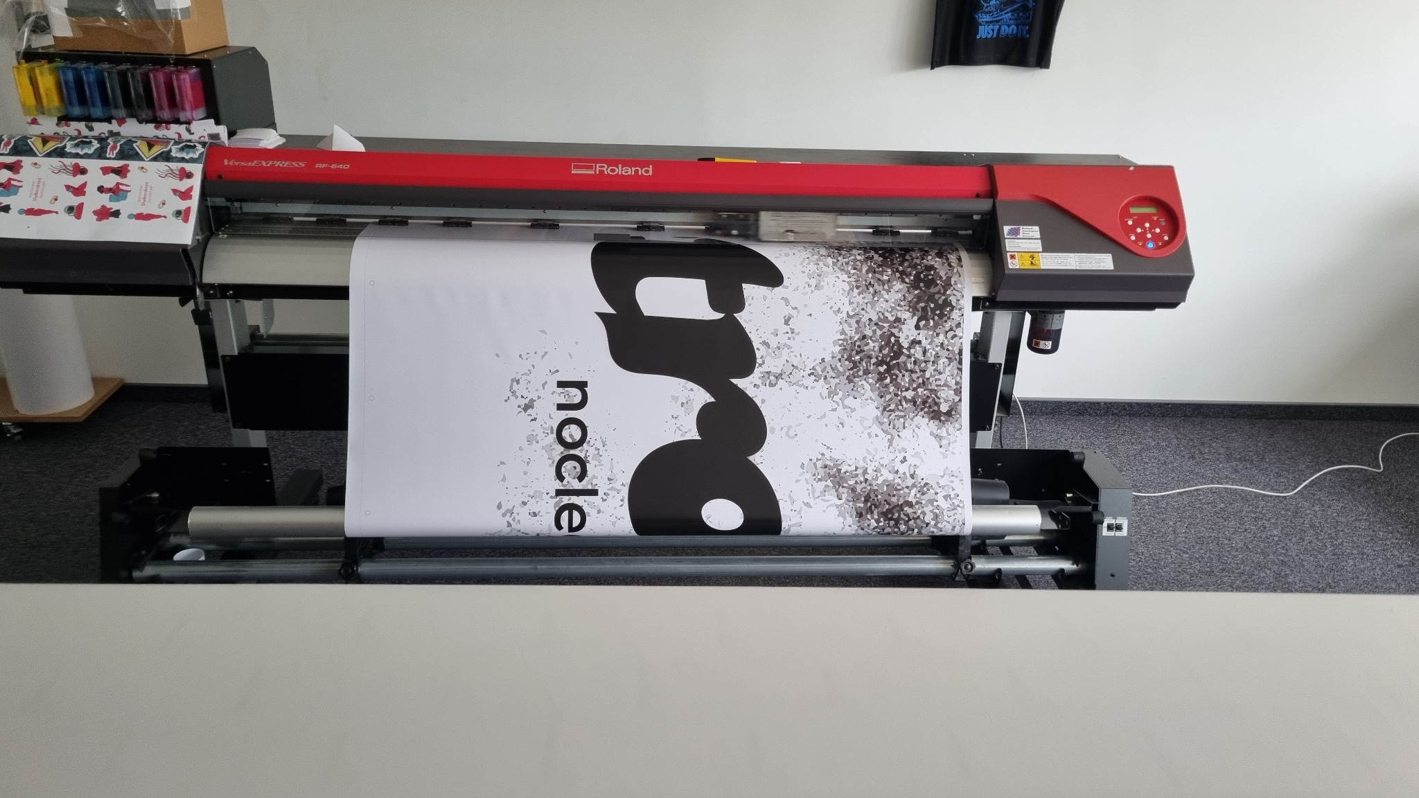 Drukarka wielkoformatowa Roland VersaExpress RF-640 w trakcie drukowania baneru z czarnym, abstrakcyjnym wzorem na białym tle. Widoczne kartridże z tuszem i panel sterowania.