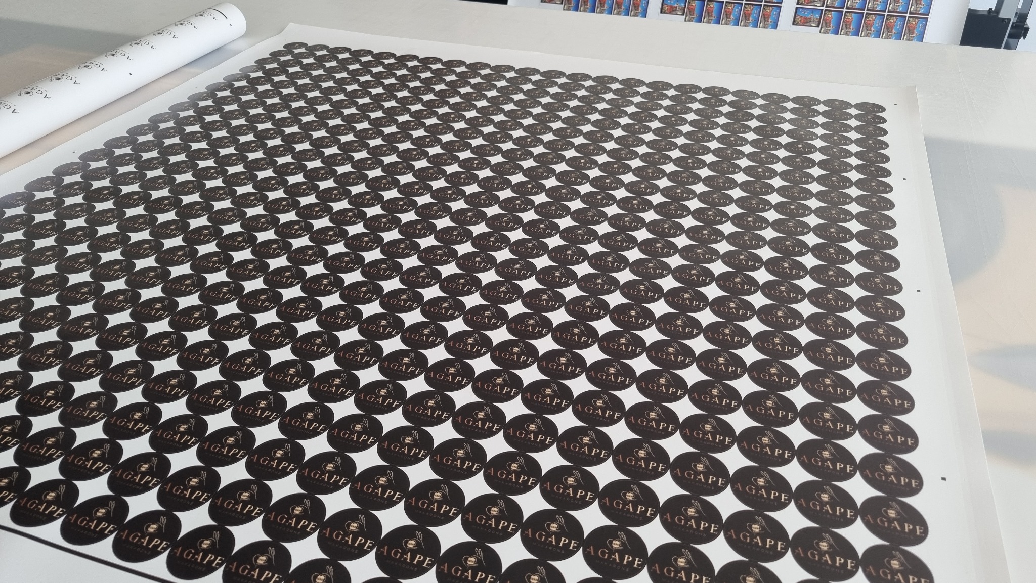 Arkusze z okrągłymi naklejkami z logo 'Agape Macarons' w kolorze czarnym i złotym, ułożone równo na białym stole, z rolką papieru w tle.