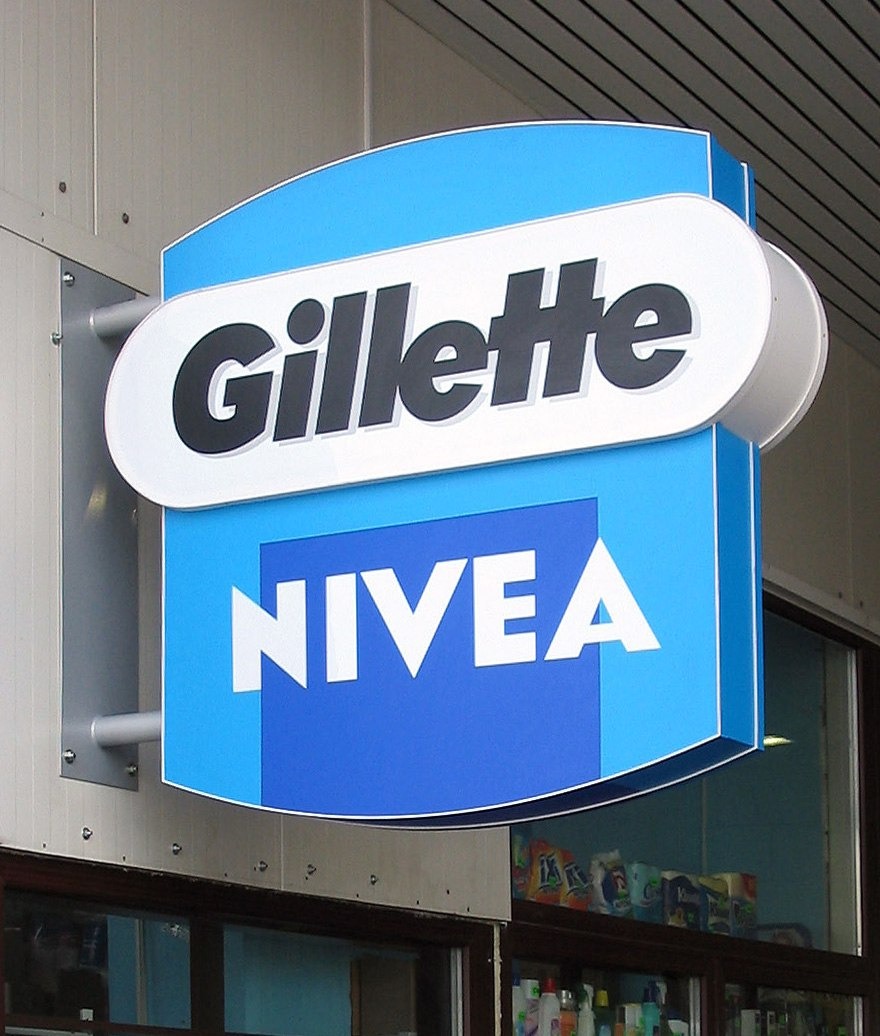 Dwustronny szyld reklamowy z logo Gillette i Nivea, zamontowany nad witryną sklepu. Szyld w odcieniach błękitu i bieli, z wyraźnymi napisami. Widoczne mocowania do ściany.