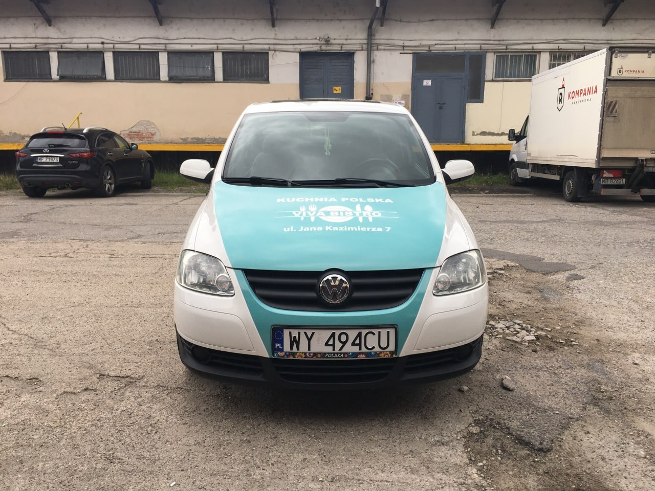 Biały Volkswagen Fox z turkusowym oklejeniem maski, prezentujący logo i adres 'Kuchnia Polska Viva Bistro'. Widok z przodu na tle magazynu i innych pojazdów.