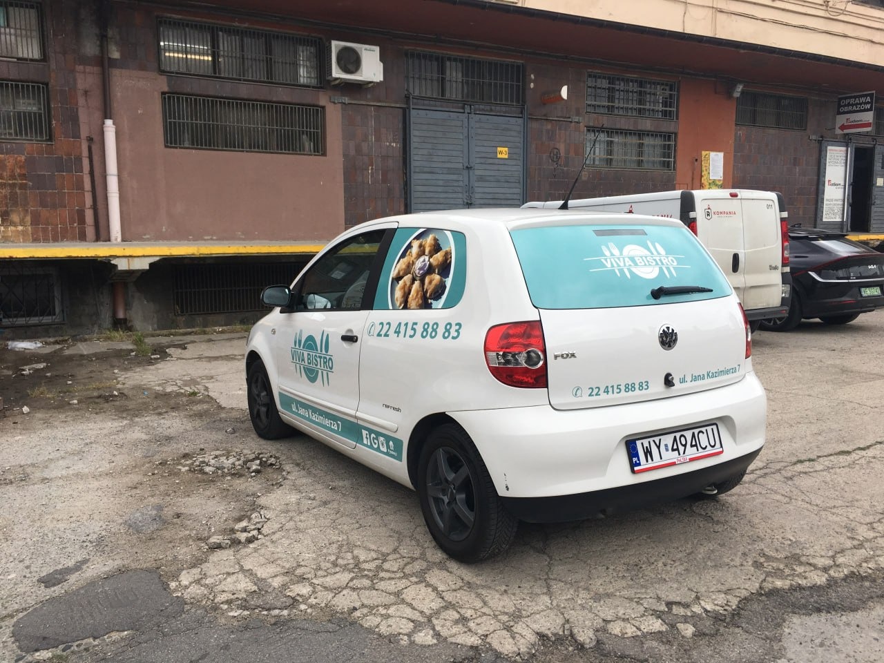 Biały samochód z reklamą Viva Bistro, numer telefonu i adres na karoserii. Grafika z jedzeniem na szybie. Auto zaparkowane na zewnątrz, widok z tyłu i boku.