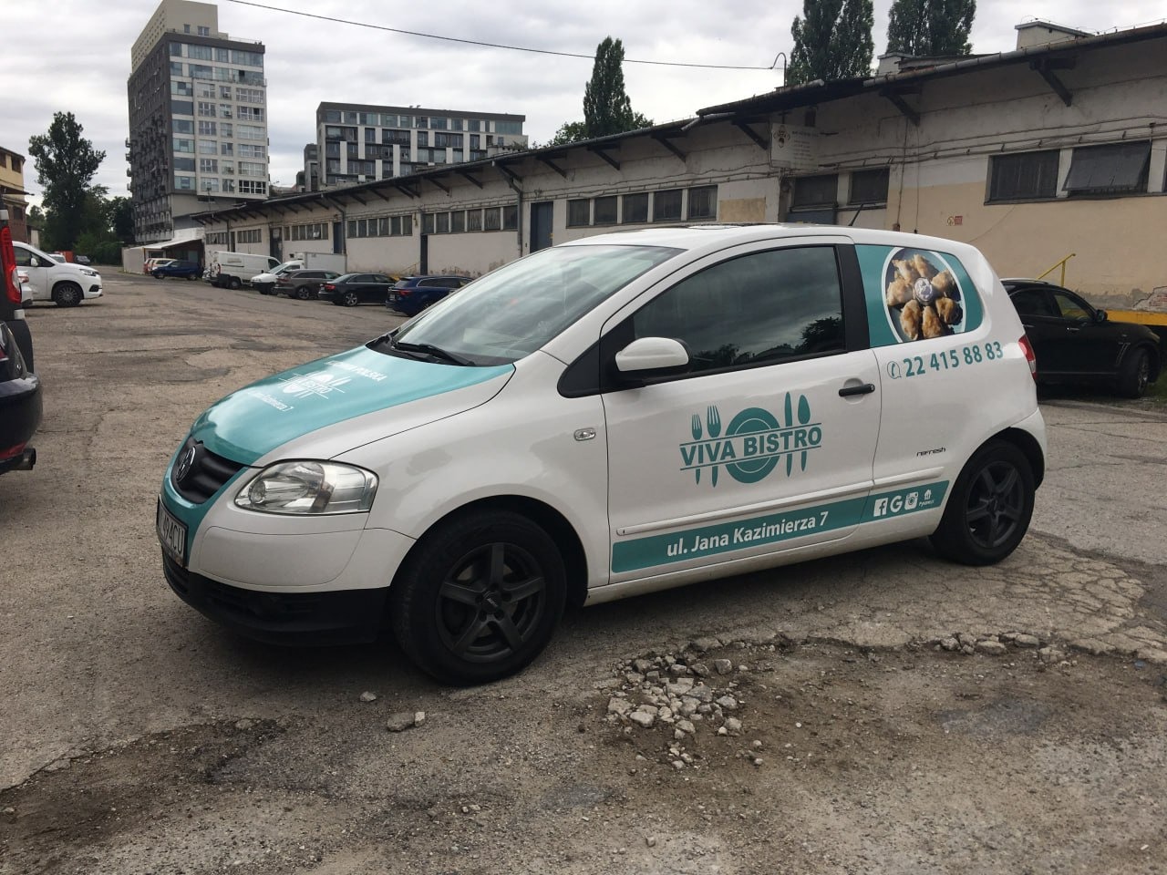 Biały samochód dostawczy z turkusowymi akcentami i logo Viva Bistro, zaparkowany na zewnątrz. Widoczne dane kontaktowe i adres na karoserii. Reklama restauracji na pojeździe.