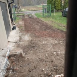 FENIX JAROSŁAW OLSZOWSKI GardenFenix Magdalena Michnik - Przygotowanie terenu pod drogę dojazdową: częściowo utwardzona nawierzchnia z gruzem, widoczne ślady prac ziemnych, fragment rozbiórki przy budynku. Pies w kadrze.