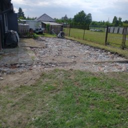 FENIX Jarosław Olszowski GardenFenix Magdalena Michnik - Fragment rozbiórki: gruz ceglany i betonowy na ziemi, widoczny ubijak wibracyjny, fragment budynku i ogrodzenie siatkowe w tle, pochmurne niebo.