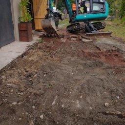 FENIX JAROSŁAW OLSZOWSKI GardenFenix Magdalena Michnik - Niebieska mini koparka w trakcie prac ziemnych na posesji. Widoczny operator w kabinie i fragment wykopu z gruzem. W tle drewniane drzwi i zieleń.