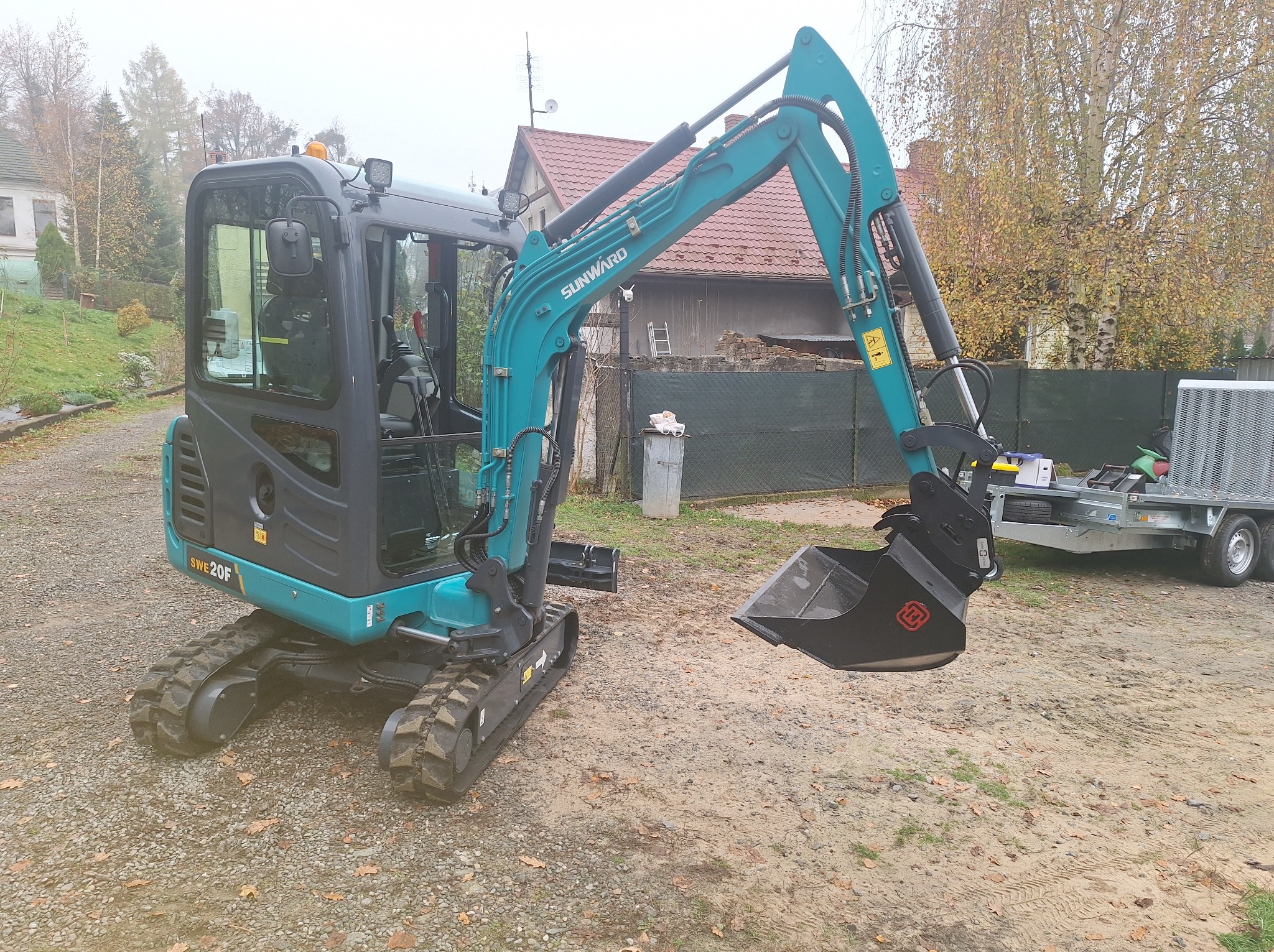 Kompaktowa, turkusowa minikoparka Sunward SWE20F na gąsienicach, z czarną łyżką, zaparkowana na placu budowy z domem w tle. Widoczna przyczepa transportowa.