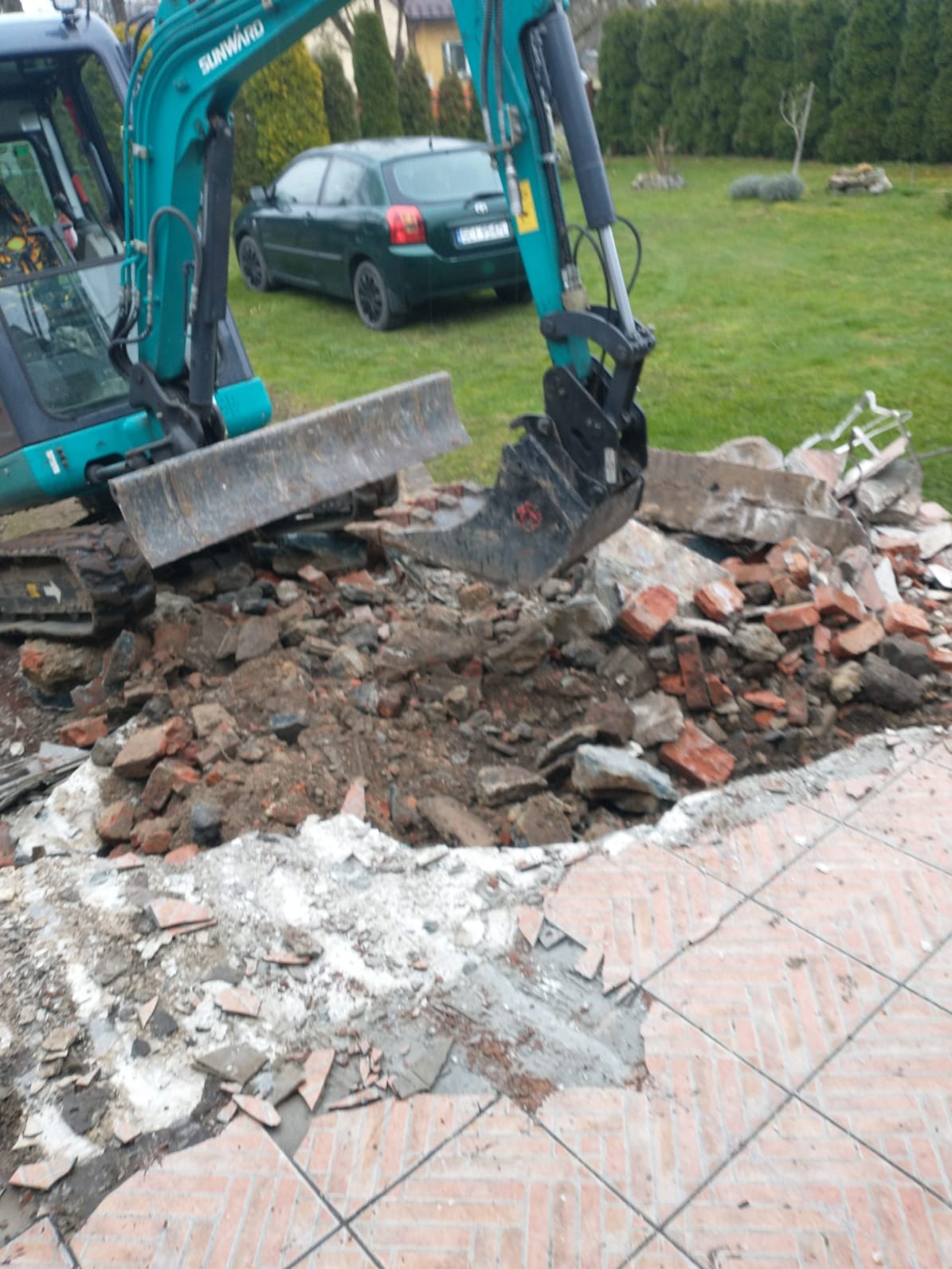 Niebieska koparka Sunward usuwa gruz i ziemię z rozbiórki chodnika z czerwonej cegły. W tle trawnik, zielony samochód i krzewy.