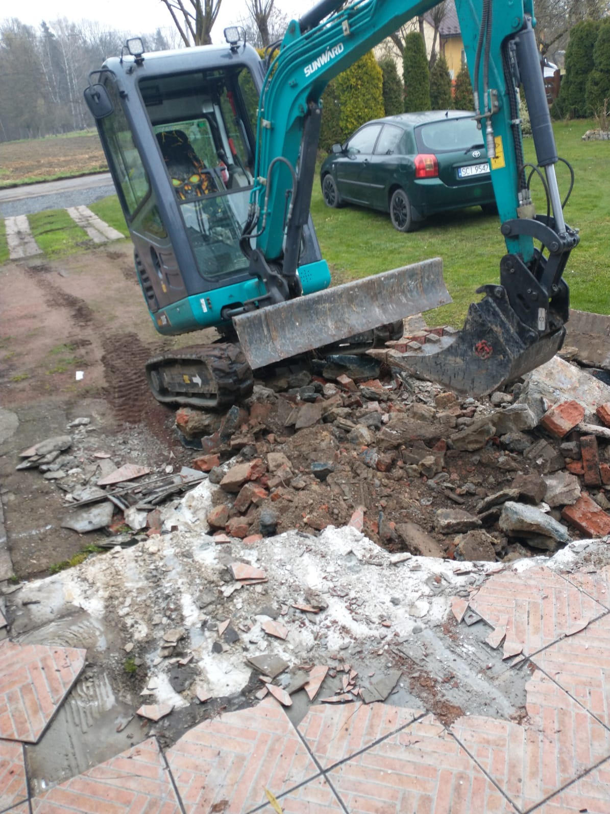 Niebiesko-szara mini koparka Sunward na gruzowisku z cegieł i płytek, obok zielony samochód osobowy i trawnik. Widoczne ślady opon na ziemi.