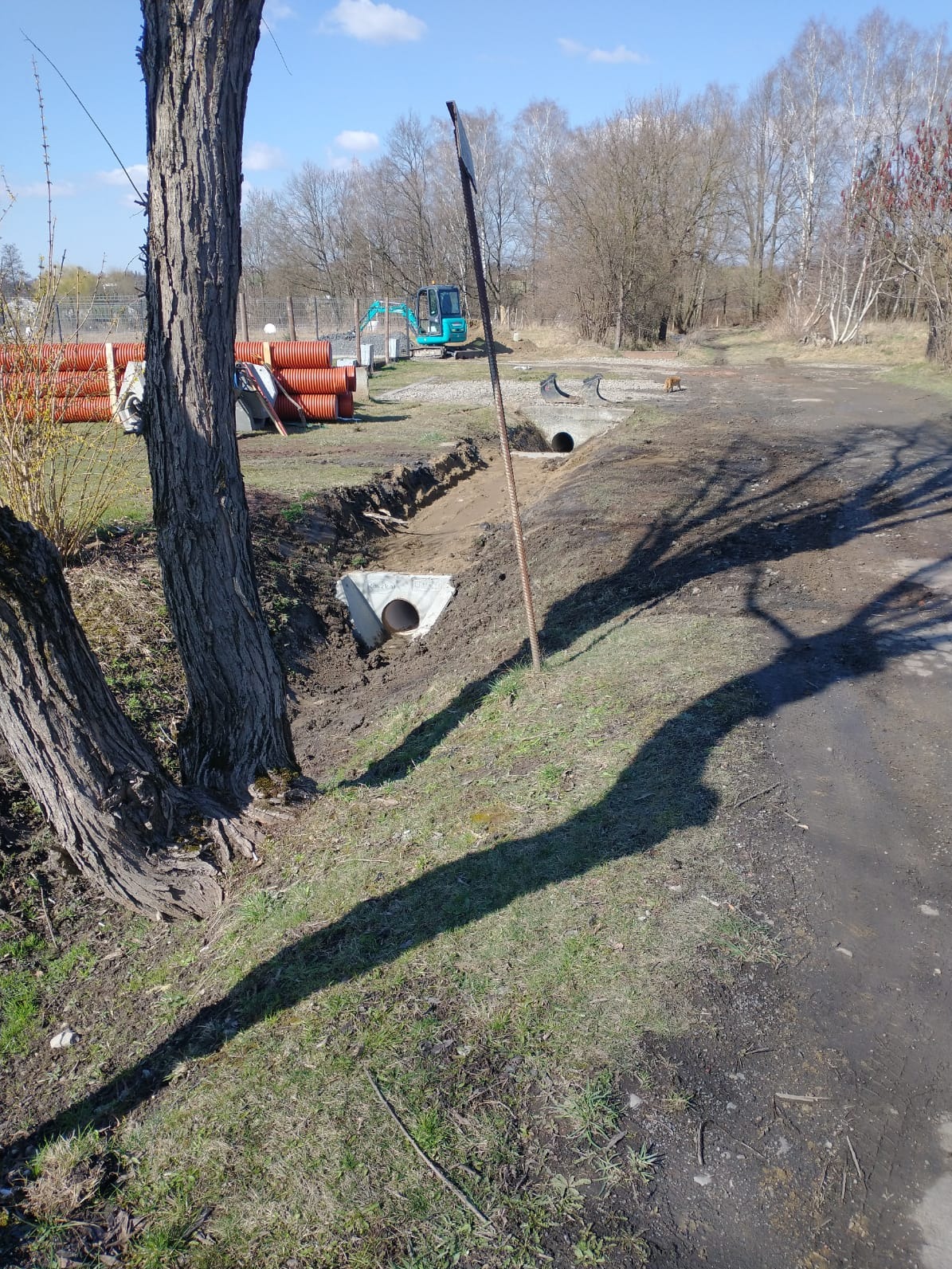 Drenaż rowu z betonowymi przepustami i rurami PCV w tle, widoczny mały, niebieski koparka. Cień drzewa na ziemi. Prace ziemne w toku, wczesna wiosna.
