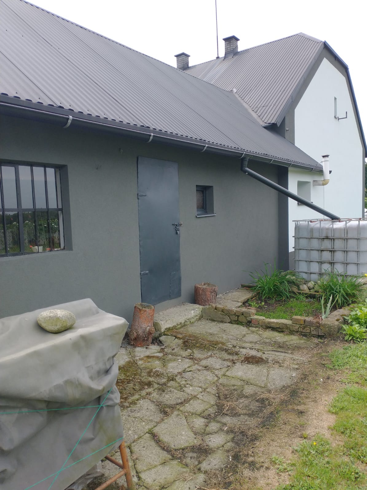 Szary budynek gospodarczy z metalowymi drzwiami i oknem z kratami. Widoczny zbiornik na deszczówkę, fragment chodnika z kostki i pniaków. Dach z blachy falistej.