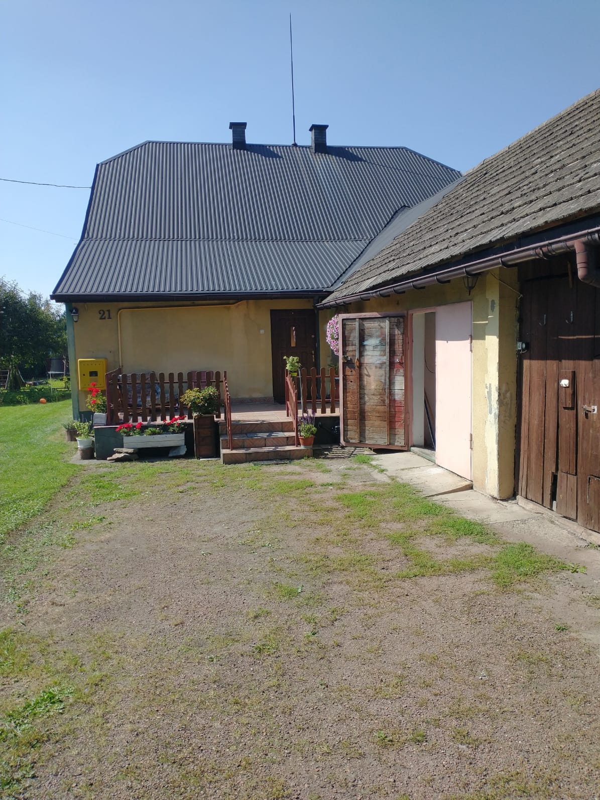 Dom z szarą blachą falistą na dachu, drewnianym ogrodzeniem i skrzynkami z czerwonymi kwiatami. Widoczny fragment podwórka z ziemią i trawą. Słoneczny dzień.