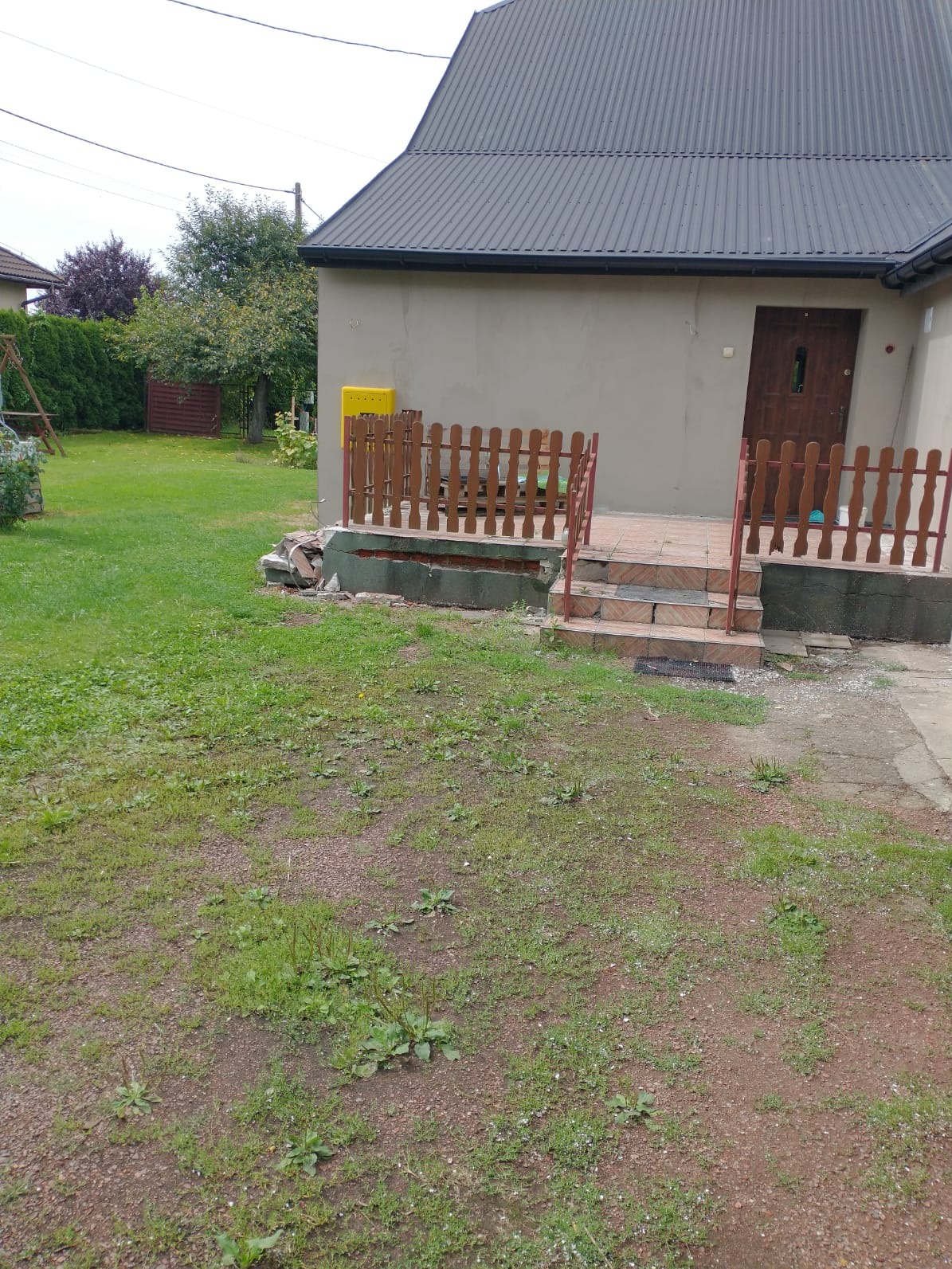 Niedokończony remont schodów wejściowych do domu z drewnianą balustradą i zniszczonymi stopniami, widok na trawnik i fragment budynku z szarą elewacją.