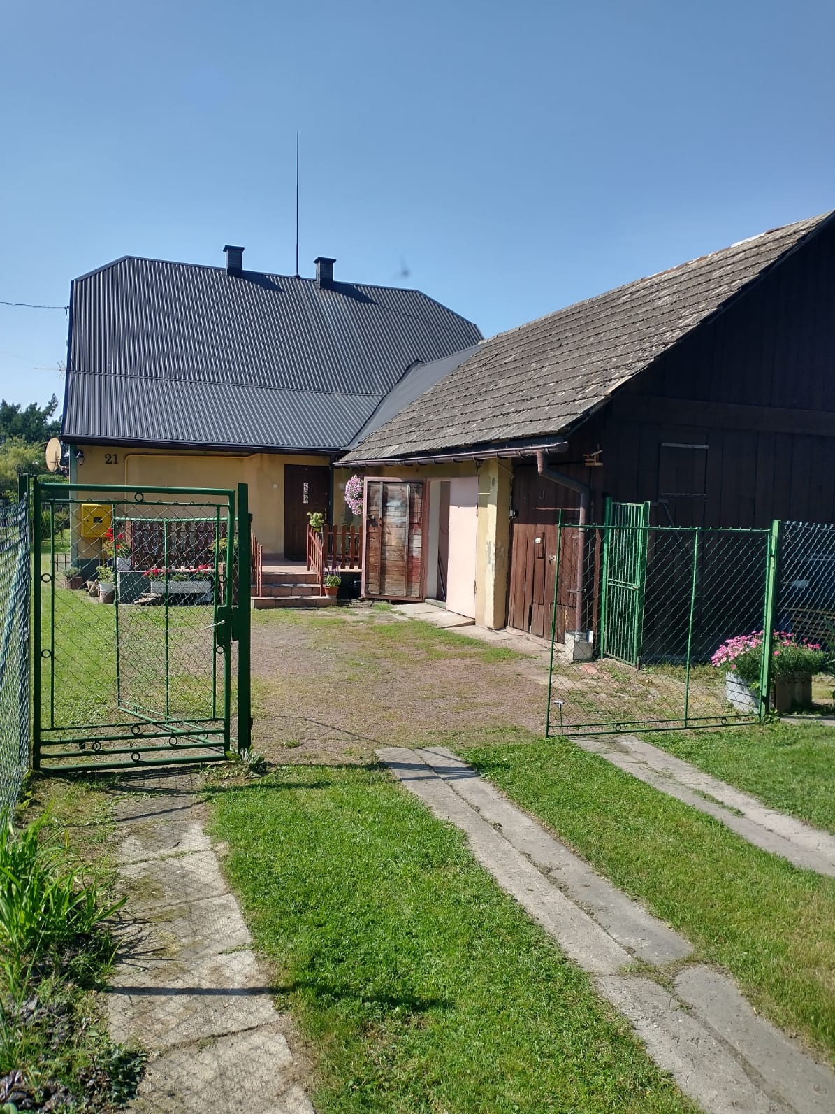 Otwarta, zielona brama z siatki na tle domu z szarą blachą falistą i drewnianej stodoły. Widoczny trawnik i betonowy podjazd. Słoneczny dzień.