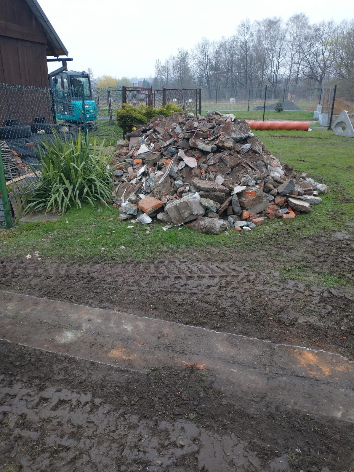 Kupa gruzu z cegłami i betonem na trawie, obok mini koparka i budynek. Ślady opon w błocie. Pomarańczowa rura w tle. Widok na teren po rozbiórce.