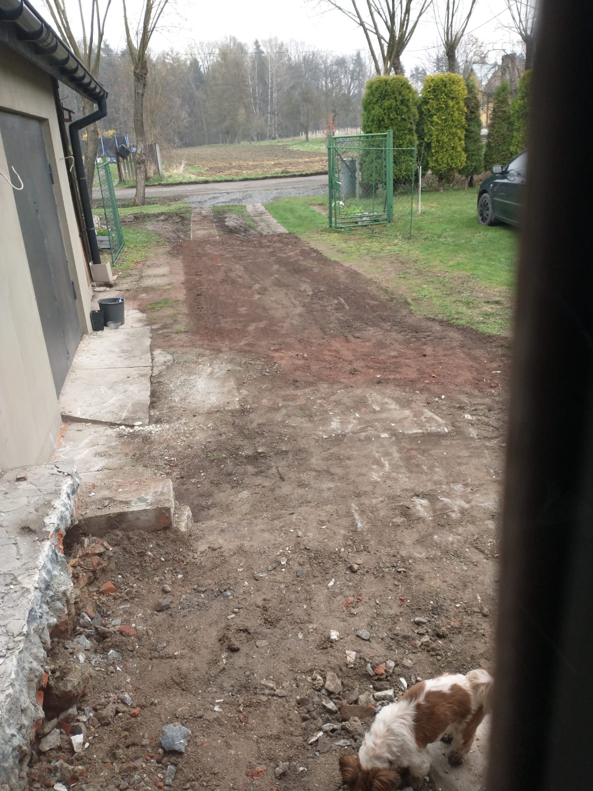 Przygotowanie terenu pod drogę dojazdową: częściowo utwardzona nawierzchnia z gruzem, widoczne ślady prac ziemnych, fragment rozbiórki przy budynku. Pies w kadrze.