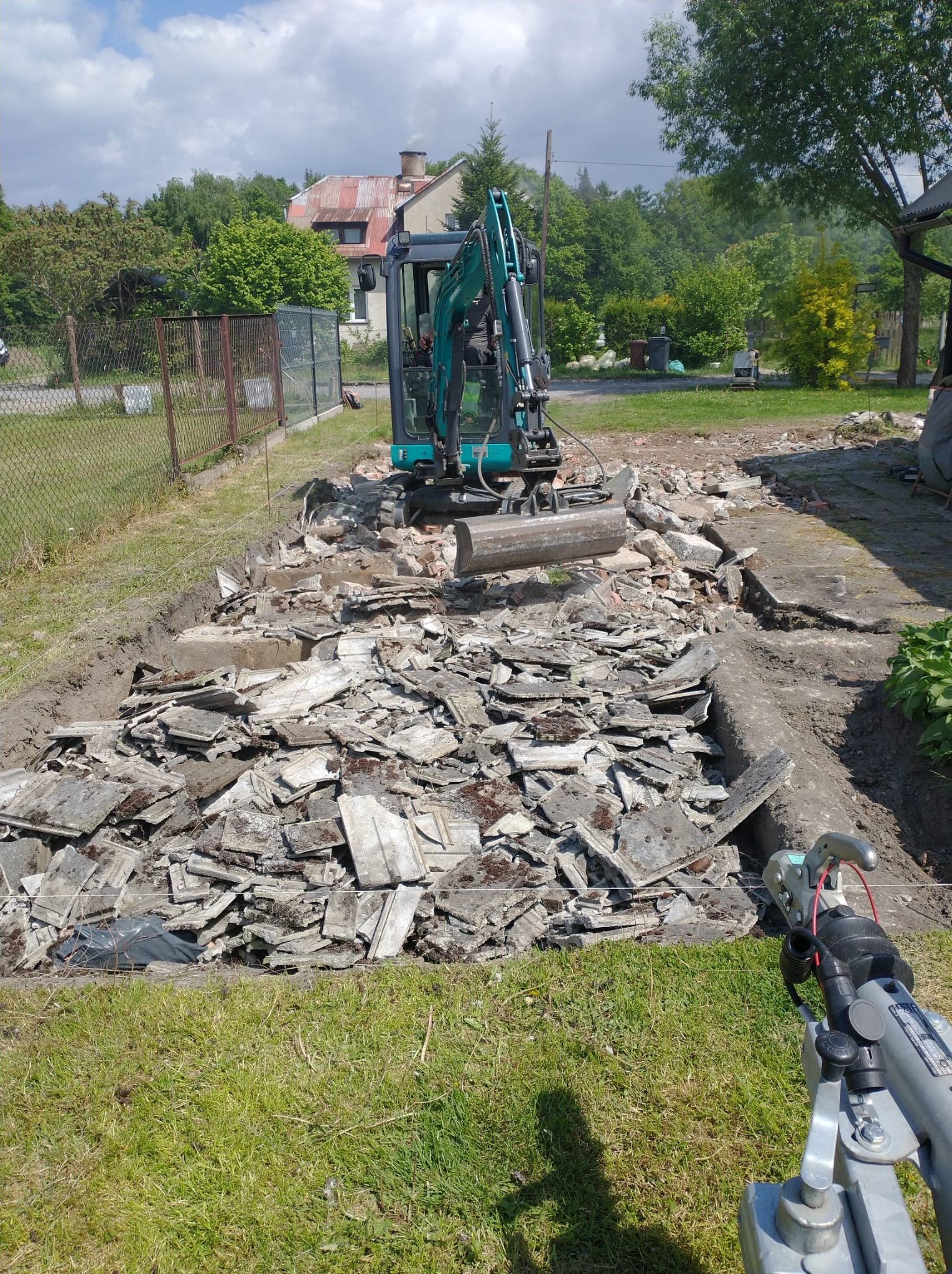 Mini koparka na terenie po rozbiórce, gruzowisko z fragmentami betonu i cegieł. Widoczne ślady wykopu i przygotowania terenu pod dalsze prace budowlane.