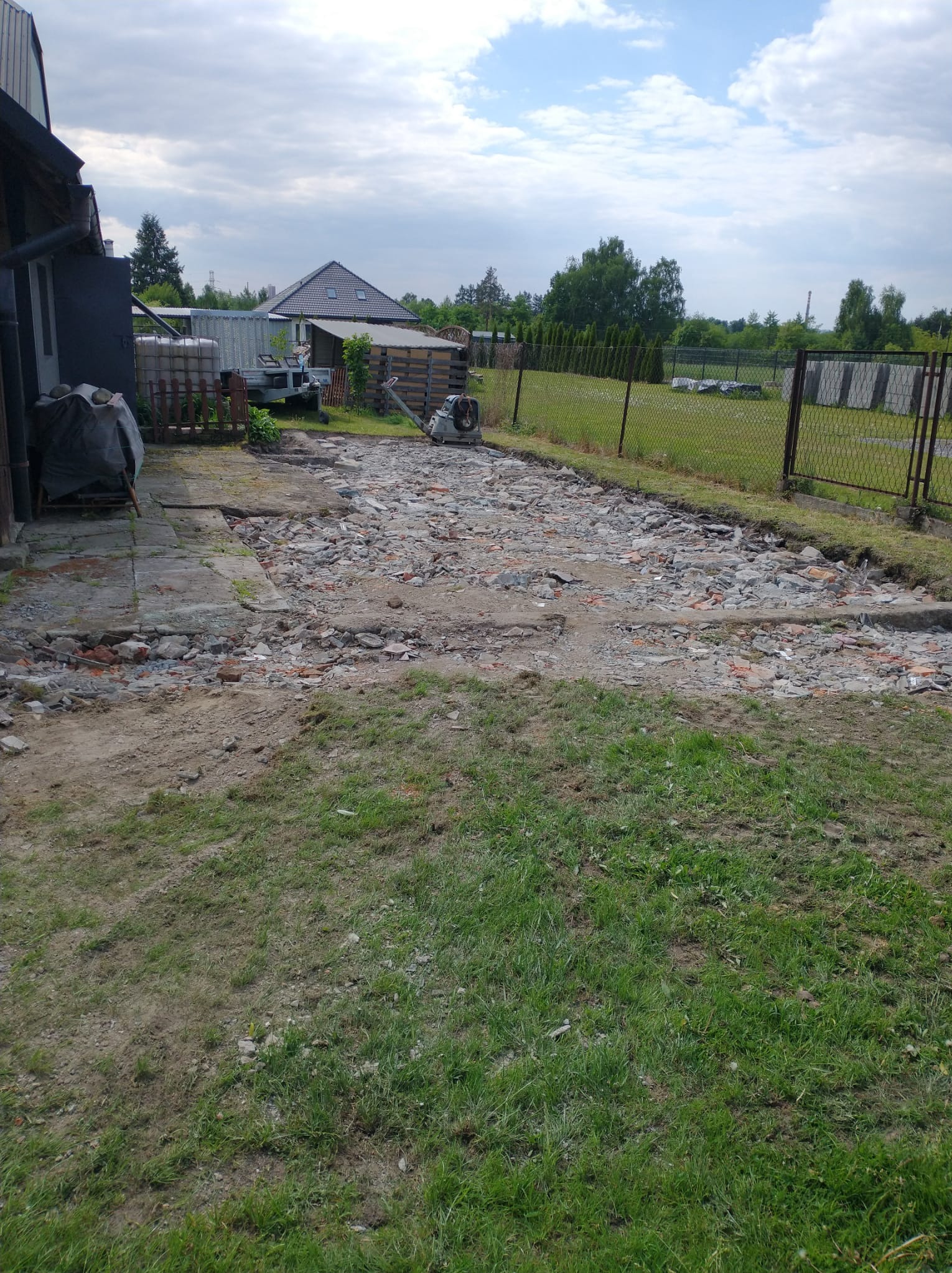 Fragment rozbiórki: gruz ceglany i betonowy na ziemi, widoczny ubijak wibracyjny, fragment budynku i ogrodzenie siatkowe w tle, pochmurne niebo.