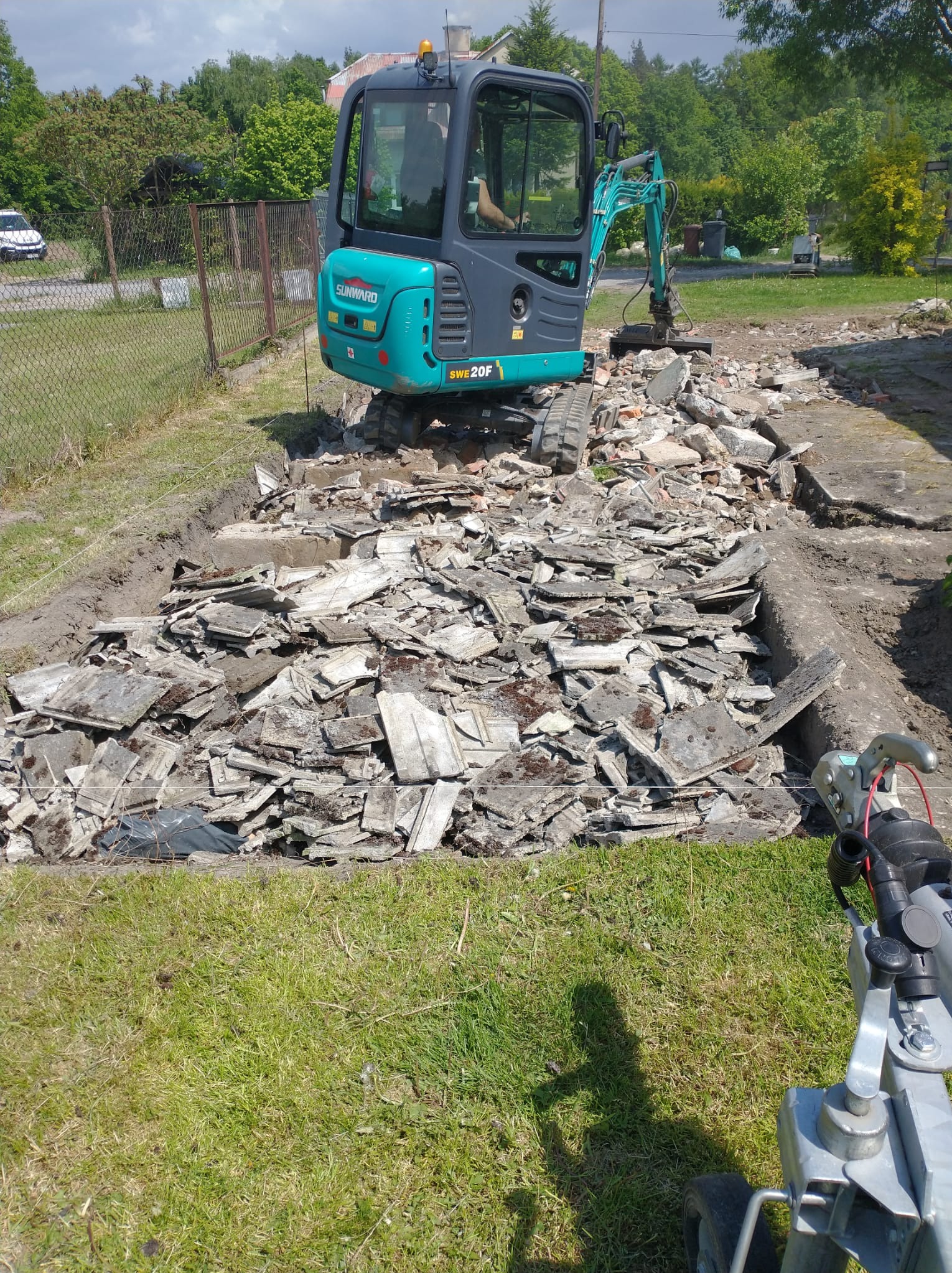 Niebiesko-szary minikoparka Sunward SWE 20F na gruzowisku z płyt, w tle zielona trawa i drzewa. Widoczny fragment przyczepy. Prace rozbiórkowe w toku.