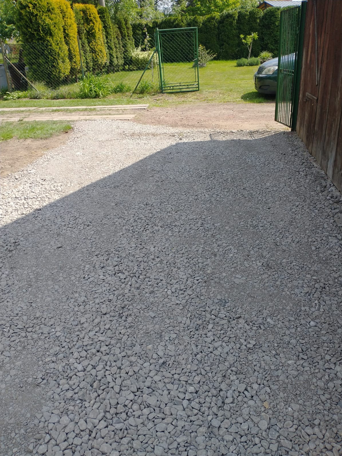 Utwardzona droga żwirowa prowadząca do zielonej bramy w ogrodzeniu, z widocznym fragmentem samochodu i zieleni w tle. Cień na żwirze.