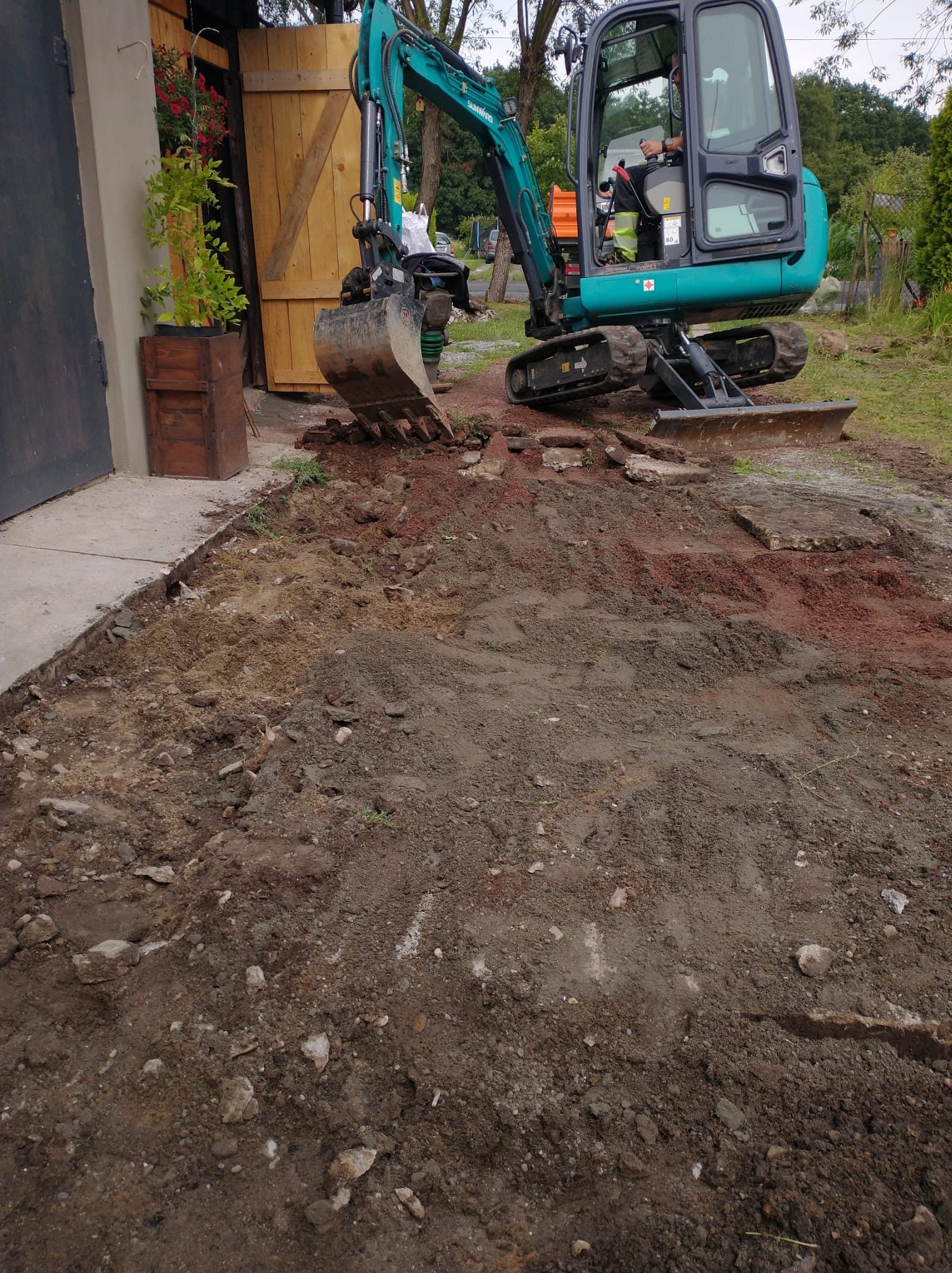 Niebieska mini koparka w trakcie prac ziemnych na posesji. Widoczny operator w kabinie i fragment wykopu z gruzem. W tle drewniane drzwi i zieleń.