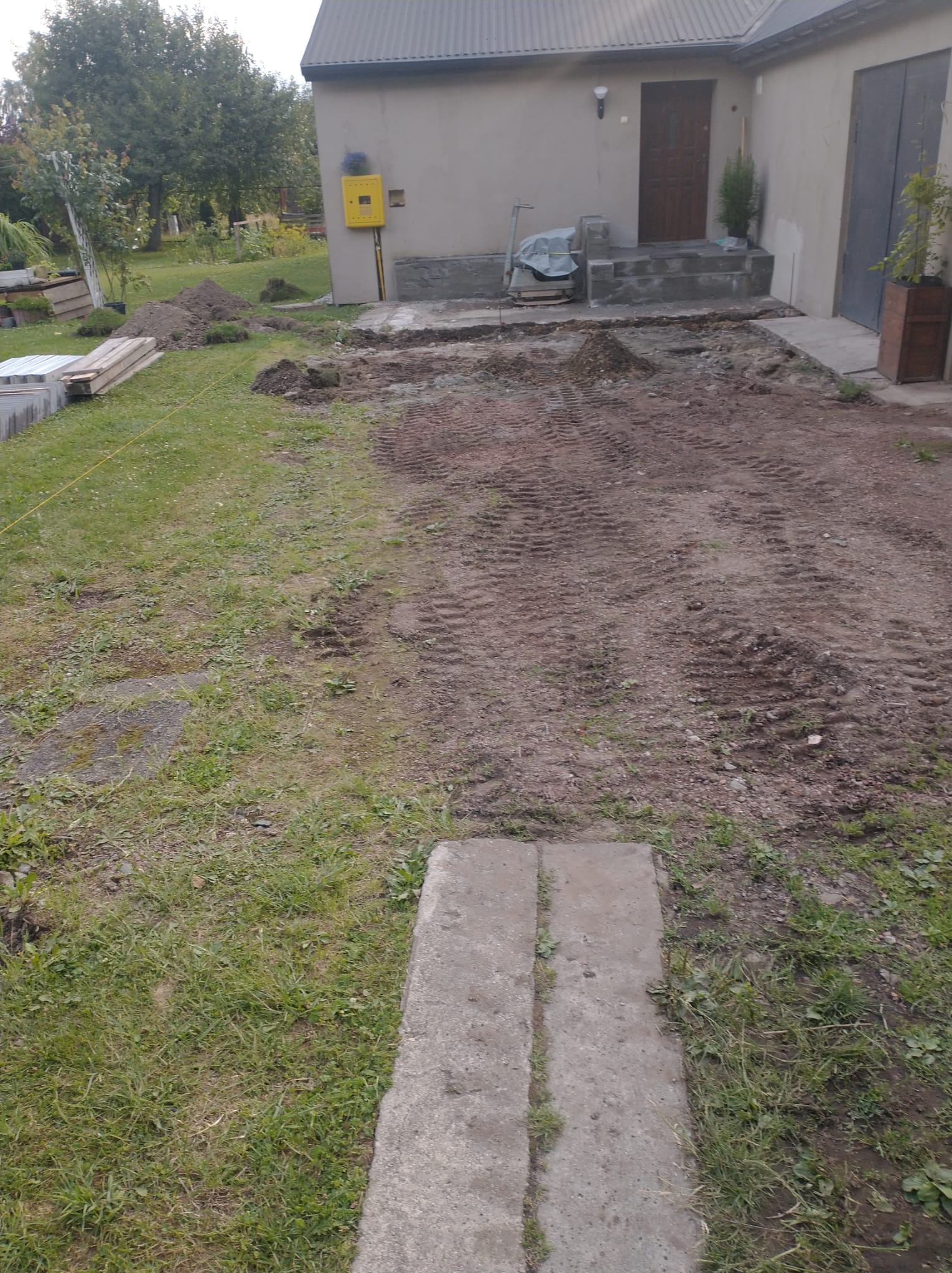Przygotowanie terenu pod budowę: wyrównana ziemia z widocznymi śladami opon, częściowo usunięta darń, materiały budowlane w tle, dom z żółtą skrzynką gazową.