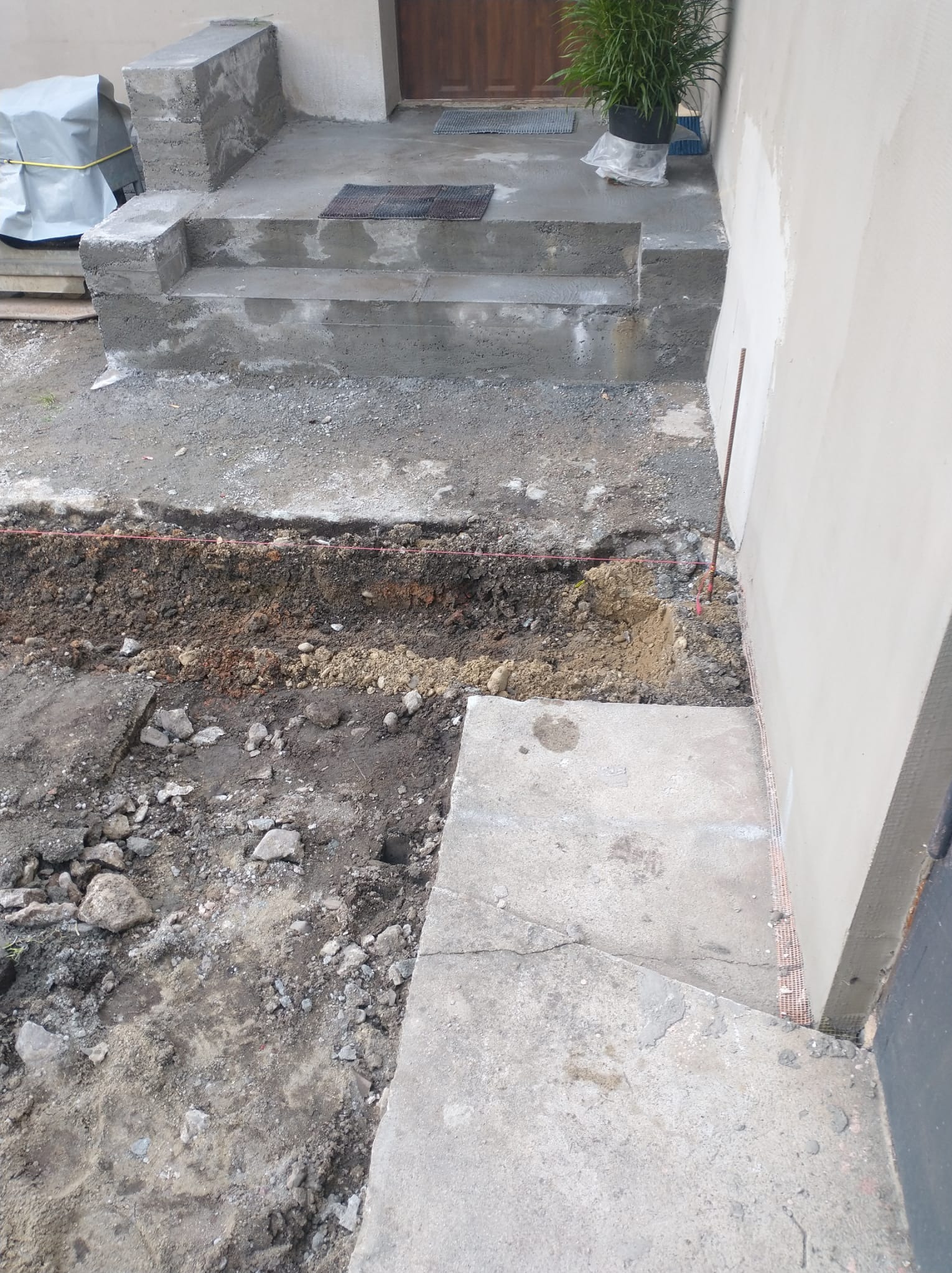 Betonowe schody wejściowe po remoncie z wykopem obok ściany budynku, widoczne narzędzia i ziemia. Prace ziemne w toku, czerwony sznurek wyznacza granice.