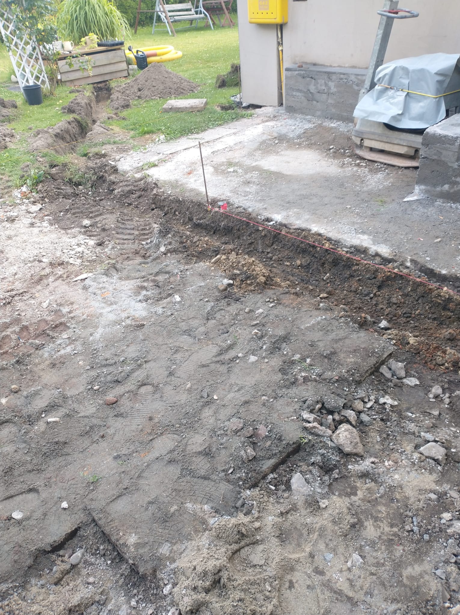 Wykopany wąski rów w ziemi, wyznaczony czerwoną linką, obok budynku z żółtą skrzynką gazową i ubijarką. Widoczne ślady opon i narzędzia ogrodowe w tle.